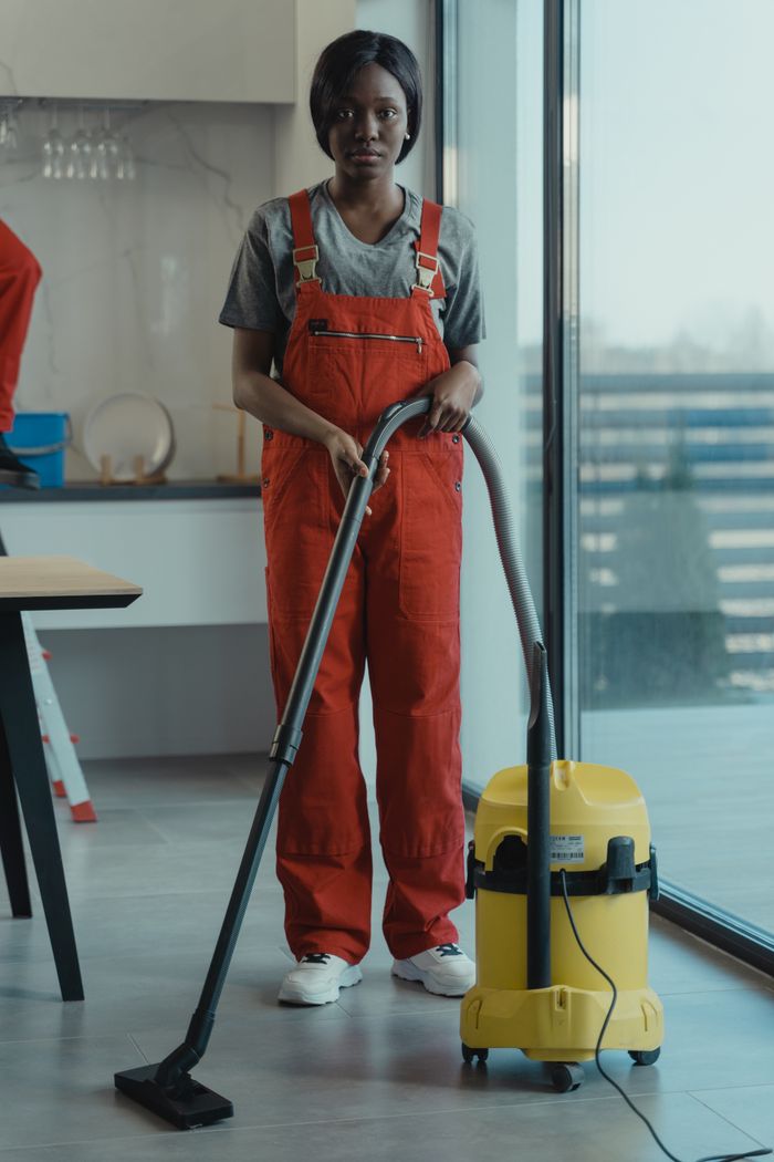 gallery-1 An adult woman in red coveralls operates a vacuum cleaner in a modern indoor setting, focusing on cleaning tasks.